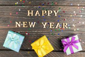 Multicolored gift boxes with tied bows. New year presents on wooden table with scattered confetti stars. Text Happy new year. Festive greeting card. Flat lay. photo