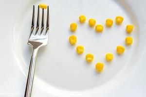 Close up of sweet corn in shape of heart and steel fork on white plate. photo
