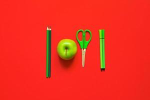 plano creativo de material escolar: bolígrafos verdes, lápices, tijeras y una manzana sobre un fondo rojo foto