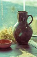 antiguo tarro de cerámica de arcilla y cuenco de arcilla sobre una mesa de madera cerca de la ventana. naturaleza muerta rústica. foto