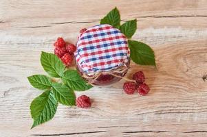 fresh raspberry jam in a glass jar on a wooden table, next to fresh raspberries with green leaves. homemade jam and preserves for winter photo