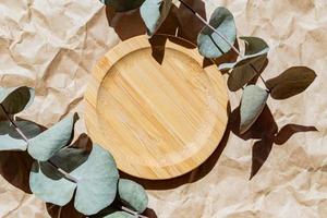 Cosmetics product advertising wooden stand. Exhibition wooden podium on paper craft background with eucalyptus dry leaves and shadows. Empty pedestal to display product packaging. Mockup. photo