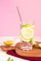 glass of homemade lemonade with lemons, mint and red paper straw on pink background. Summer refreshing beverage. photo