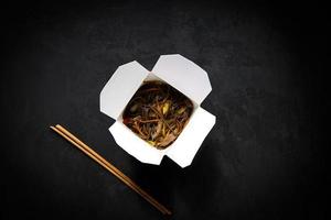 Asian restaurant food delivery. Soba noodles with meat, vegetables and soy sauce in white take-out paper box on black background with chopsticks, top view, copy space photo