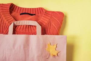 Orange woman warm sweater in paper craft package on yellow background with autumn maple leaves decoration. Clothing sale and black friday concept.  Top view. photo