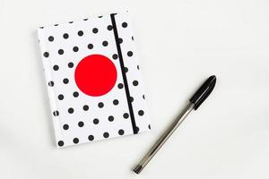 Black and white polka dot note book with red circle  on the cover and black pen on white table. top view, minimal flat lay photo