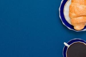 top view of cup of black coffe and croissant on a plate on blue background with copy space. Morning breakfast in french style. photo