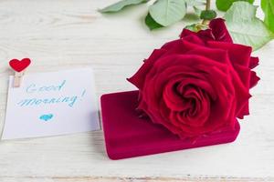 caja de regalo de joyería y hermosa rosa roja sobre fondo de madera. deseando buenos días en papel blanco foto