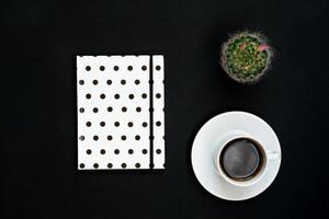 Cup of black coffee, notebook and blooming cactus on black background, Top view, copy space. photo