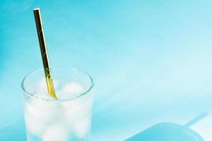 Close up of pure water with ice and paper straw in transparent glass and sun glare on blue background. photo