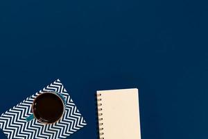 top view of cup of black coffe and clean notebook on blue background with copy space. photo