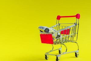 Toy car in small shopping cart on yellow background. Car rent and consumering concept with copy space photo