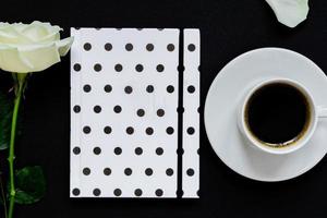 Cup of black coffee, notebook and white rose on black background, Top view, copy space. photo