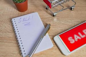 close up view of office desk with white smartphone with sale text, note and small shopping cart. Technology business online shopping concept photo