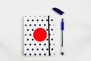 Black and white polka dot note book with red circle  on the cover and blue pen on white table. top view, minimal flat lay photo