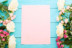 Frame of colorful roses on blue wooden background with paper card and copy space. Top view and selective focus. Mock up photo