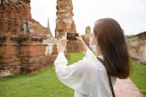 young beautiful woman wearing protective mask traveling and taking photo at thai historical Park, Holidays and cultural tourism concept.