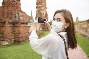 young beautiful woman wearing protective mask traveling and taking photo at thai historical Park, Holidays and cultural tourism concept.