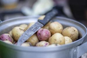 cacerola de acero inoxidable con patatas nuevas crudas y un cuchillo de cocina. papas sin pelar. primer plano de papas de piel amarilla y roja. pelar patatas en la cocina sobre una mesa rústica de madera. foto