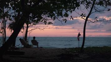 Paare sitzen und beobachten den Sonnenuntergang am Meer. Silhouetten von Frauen und Männern, die auf einer Bank sitzen und den ruhigen Sonnenuntergang über dem Ozean beobachten. entspannungs- und urlaubsreisekonzept video