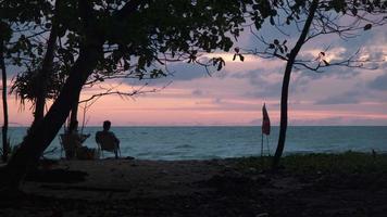coppie sedersi e orologio il tramonto di il mare. sagome di donne e uomini seduta su un' panchina Guardando il sereno tramonto al di sopra di il oceano. rilassamento e vacanza viaggio concetto video