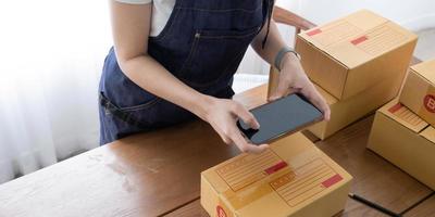 Hands of Asian woman entrepreneur holding smartphone with cardboard boxes, computer laptop, online selling equipments. Packing, business and technology concept. Top view, copy space photo