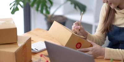 retrato, empresaria, joven asiática, empresa independiente, pyme, compras en línea, trabajo en computadora portátil con caja de paquetes en la mesa de la oficina en casa, negocio en línea y concepto de entrega. foto