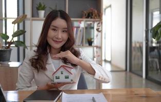 Miniature house in the hands of an Asian woman real estate agent home loan working at the office. Looking at the camera. photo