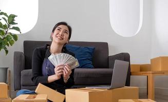 Young woman freelancer working and holding money with cardboard box  at home - SME business online and delivery concept photo