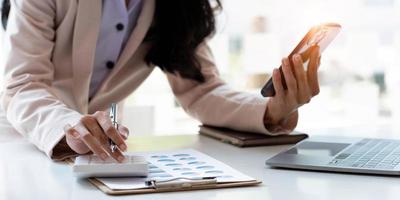 mano de mujer de negocios o contable usando calculadora, trabajando en un teléfono inteligente móvil y una computadora portátil con papel gráfico de datos documentales sobre la mesa en la oficina doméstica moderna. concepto de marketing en línea. foto