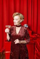 mature stylish elegant woman with glass of sparkling wine with presents on red background. Party, fashion, celebration, anti age concept photo