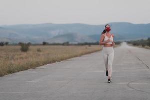 Determined fitness woman in short clothes wearing red protective face mask running outdoors in the city during coronavirus outbreak. Covid 19 and physical jogging activity sport and fitness. photo