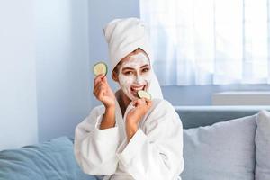 Beautiful young woman with facial mask on her face holding slices of fresh cucumber. Skin care and treatment, spa, natural beauty and cosmetology concept. photo