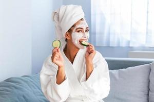 Beautiful young woman with facial mask on her face holding slices of fresh cucumber. Skin care and treatment, spa, natural beauty and cosmetology concept. photo