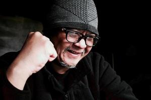 Bearded Asian man in black jacket and knitted hat. Working in a dark room and being happy and successful photo