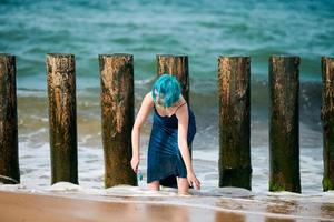 Beautiful blue-haired woman performance artist in blue dress standing on beach holding paint brush photo