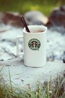 taza de café starbucks colocada sobre rocas alrededor de una fogata de camping con fondo borroso, tonos modernos. foto