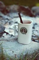 taza de café starbucks colocada sobre rocas alrededor de una fogata de camping con fondo borroso, tonos modernos. foto