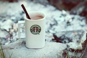 taza de café starbucks colocada sobre rocas alrededor de una fogata de camping con fondo borroso, tonos modernos. foto