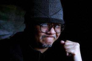 Bearded Asian man in black jacket and knitted hat. Working in a dark room and feeling successful photo