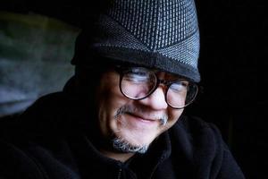 hombre asiático barbudo con chaqueta negra y gorro de punto. trabajando en una habitación oscura y sonriendo felizmente foto