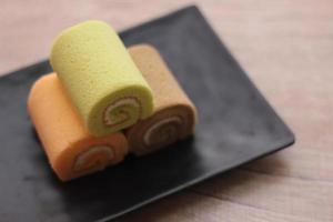 3 Swiss rolls served in a black plate placed on a wooden table. photo