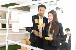 businesspeople office worker reading sticky notes on planning glass board. Business team pointing on keywords in sticky notes and business report to brainstorming about planning strategy of business. photo