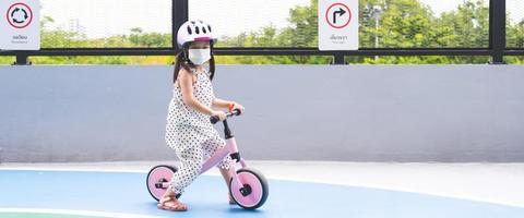 Cute Asian girl is playing on bicycle with her legs plowed on ground. Children wear helmets while practicing. Behind kid is traffic sign written in Thai and English that says Roundabout and Turn right