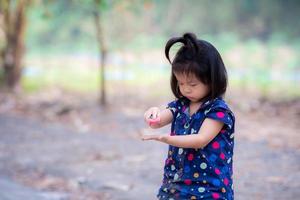 una linda niña asiática está jugando con un camión de juguete rojo. hay manchas de tierra en su ropa de jugar. niño jugando en el campo. niño tiene tres años. foto