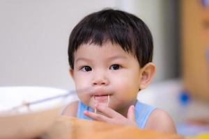 A 2-year-old child boy uses his hand to pick up instant noodles. Asian baby boy put a line of instant noodles in his mouth and looked at the camera. Son is eating food in the house. photo