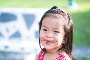 Candid image baby 3-4 years old. Cute little Asian girl is licking chocolate smeared all over her lips and cheeks. Face children enjoy dessert in afternoon. Summer or spring season. Child sweet smile. photo