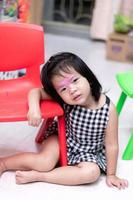 Asian girl is leaning on red chair. Child use makeup to imitate adult. The colors on face of baby that is difficult to wash off. Happy children sweet smiles. Adorable kids aged 3-4 year. On cloudy day photo