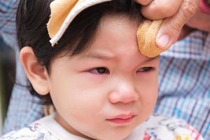 Boy is crying out of pain. Child bumps the head against solid. Baby is bulging. Adult is wrapping ice cube in cold compress on child. Closeup, crying son face from injury caused by home accident. photo