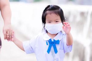 Student wearing white face mask walks hand in hand with her family to school in new happy way of life. New Normal. Adorable child girl had bright eyes. Cheerful kid aged 3 years old. photo